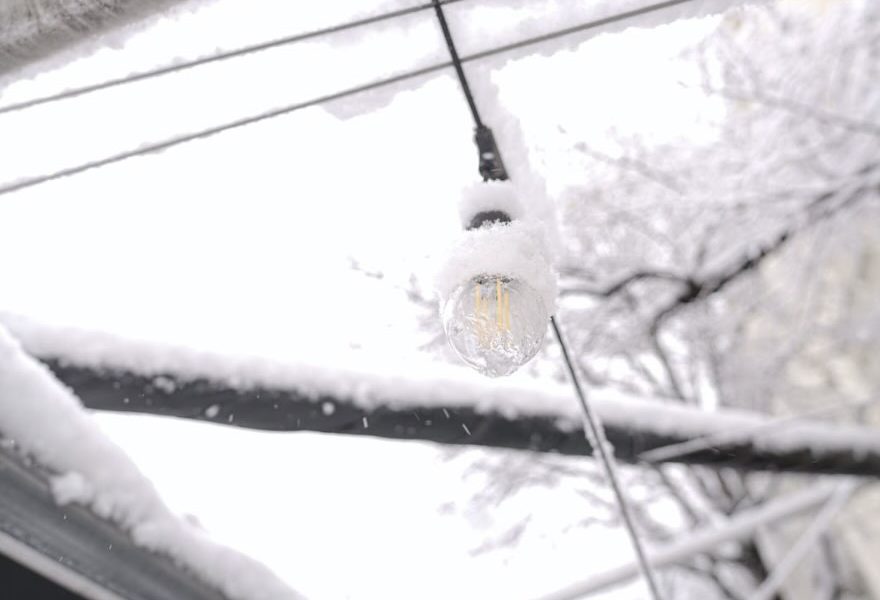 A light bulb hanging from a wire in the snow. The bulb illuminates the wintry scene, creating a contrast between warmth and coldness.